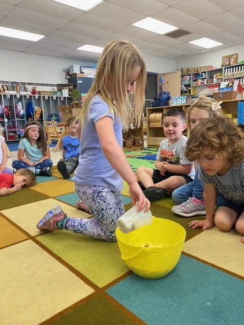 A little girl is pouring a cup of food into a bowl to make a class trail mix as kids look on.