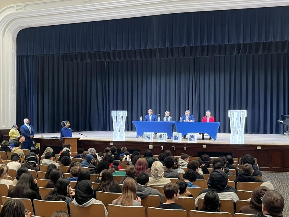 Individuals on a stage sitting behind blue tables while students sit in the audience. 