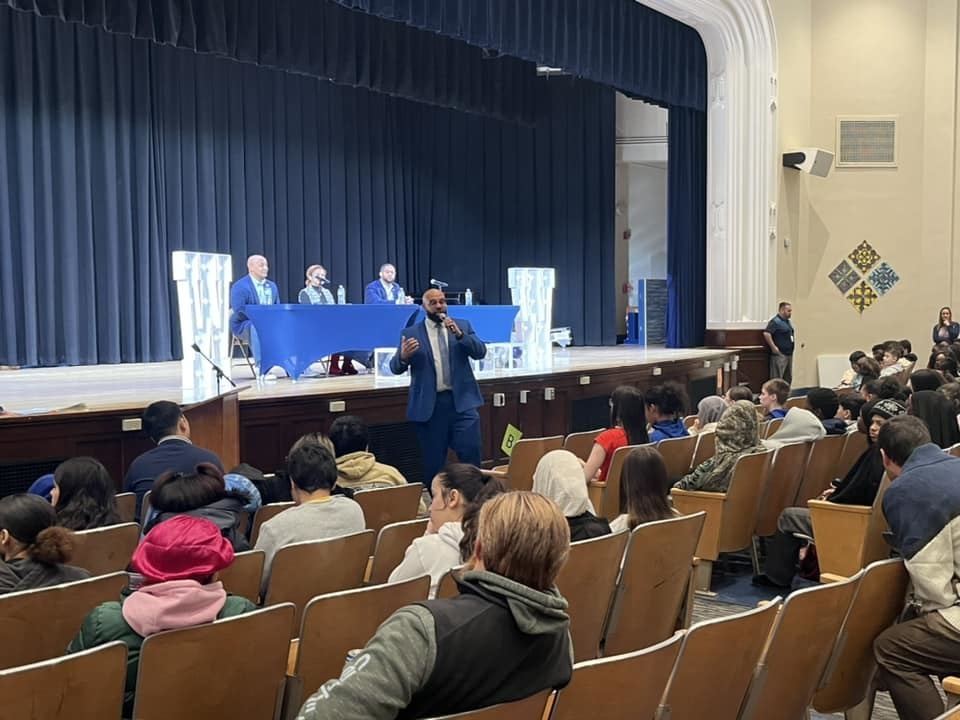 Individuals on a stage sitting behind blue tables while students sit in the audience. 