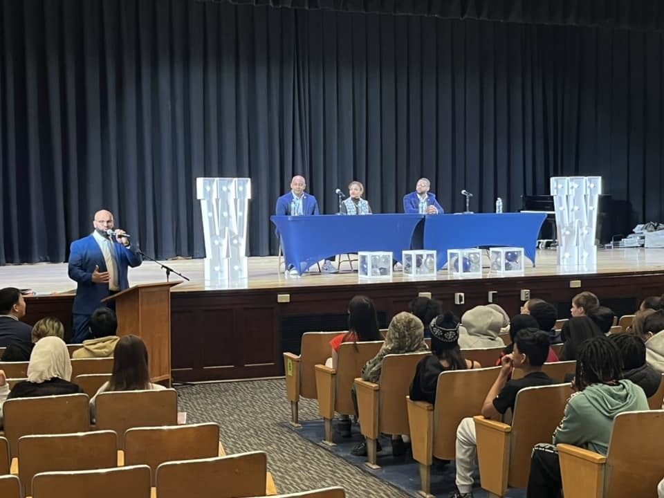 Individuals on a stage sitting behind blue tables while students sit in the audience. 