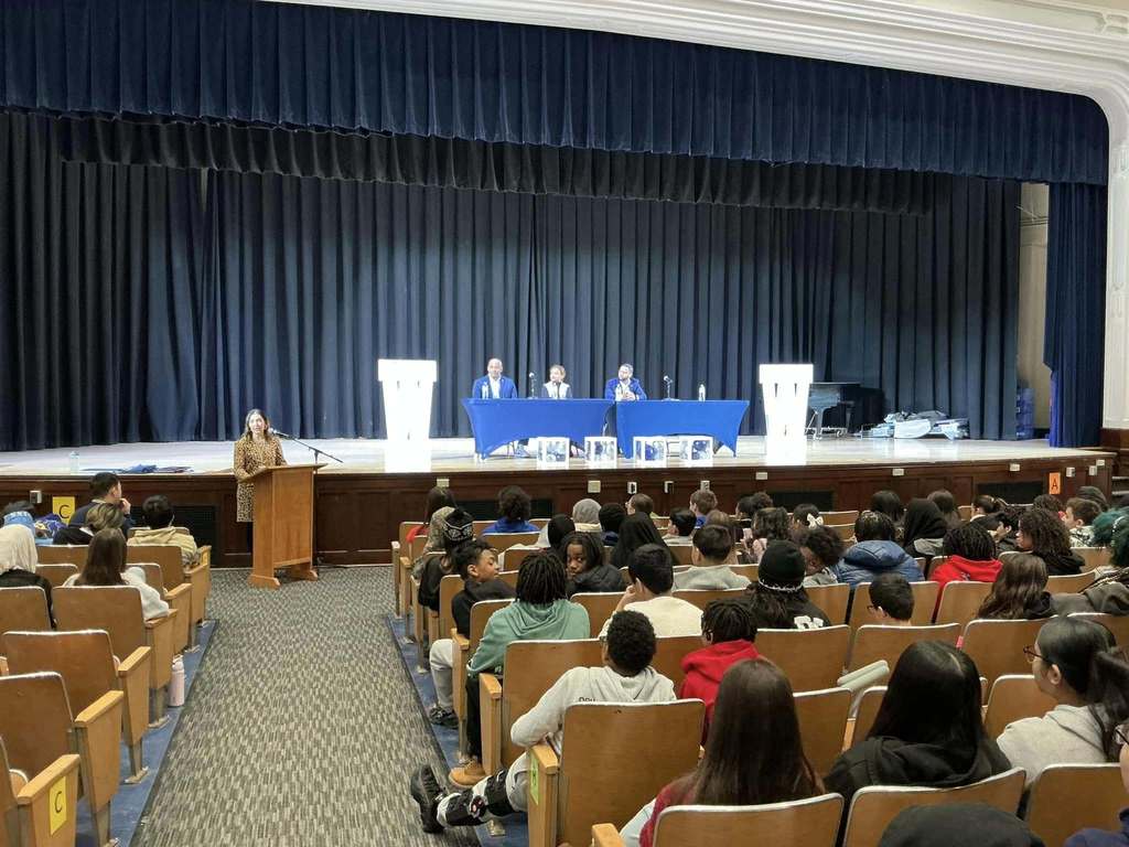 Individuals on a stage sitting behind blue tables while students sit in the audience. 