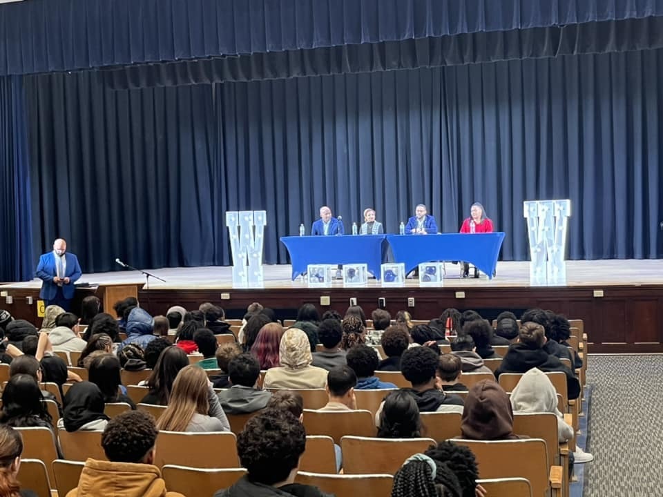 Individuals on a stage sitting behind blue tables while students sit in the audience. 