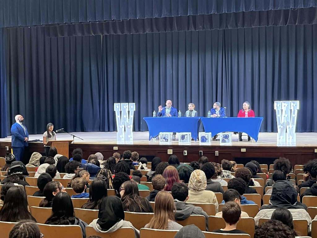 Individuals on a stage sitting behind blue tables while students sit in the audience. 