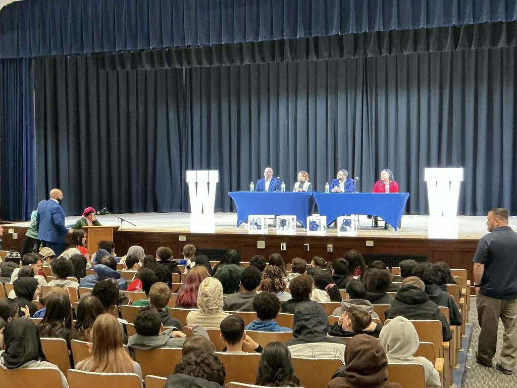 Individuals on a stage sitting behind blue tables while students sit in the audience. 
