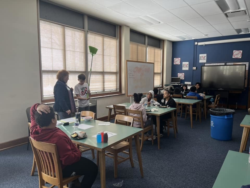 Kids sitting in a classroom while an adult shows a child how to do an experiment. 