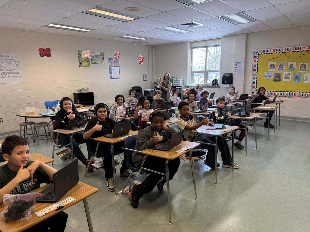 Students in a classroom giving a thumbs up and their laptops are open in front of them.