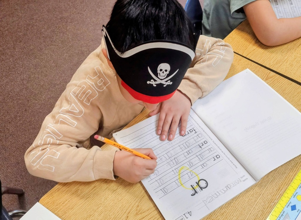 young kid in a tan shirt writing in class with a pirate hat on his head.
