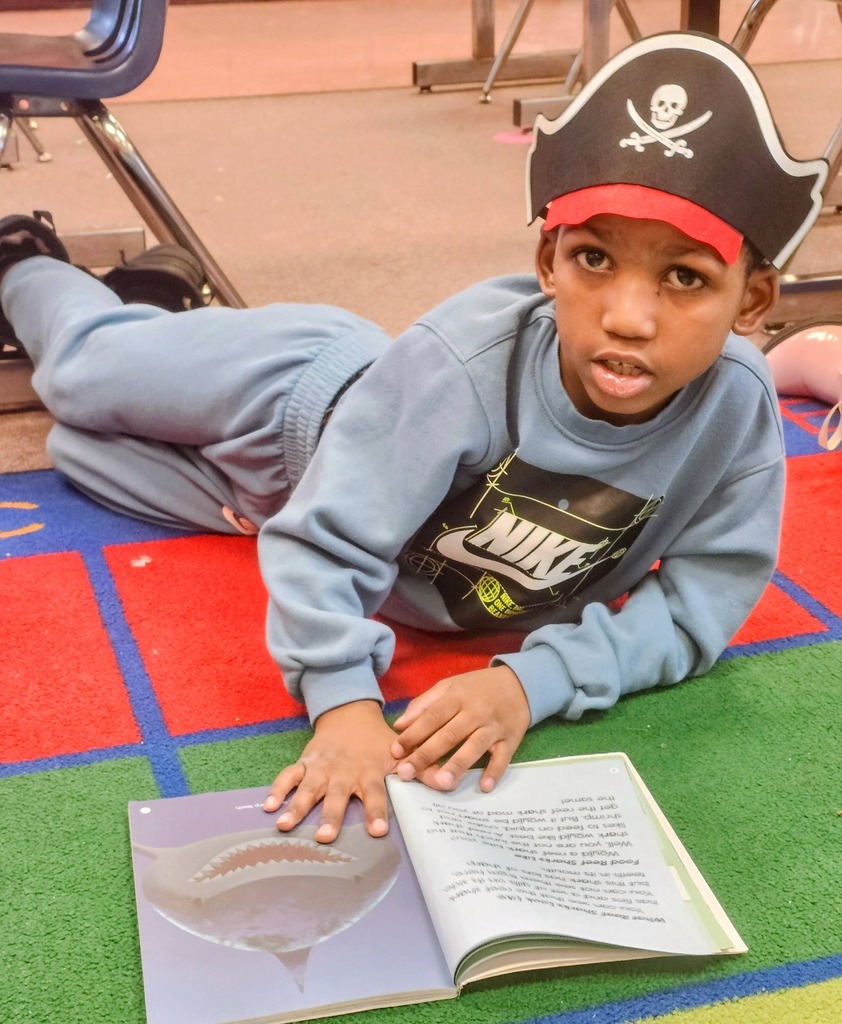 A kid reading on a colorful floor in class with a pirate hat on his head.