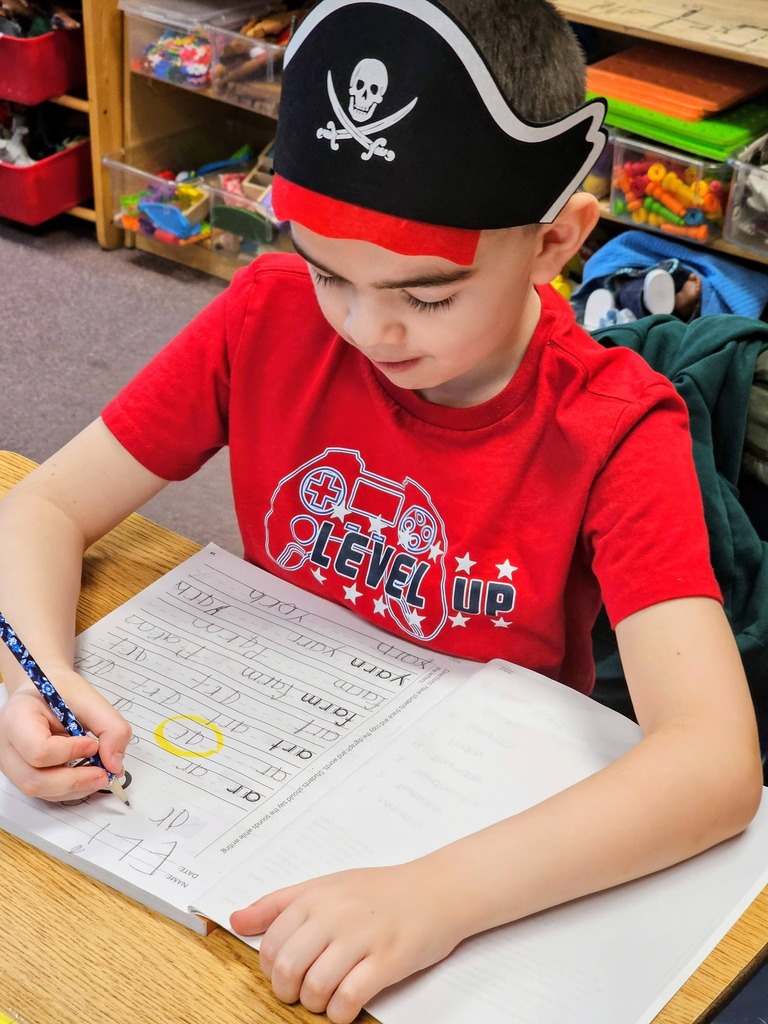 Young boy in a red shirt writing in class with a pirate hat on his head.
