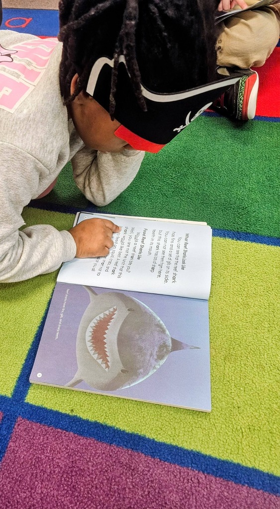 A kid reading on a colorful floor in class with a pirate hat on his head.