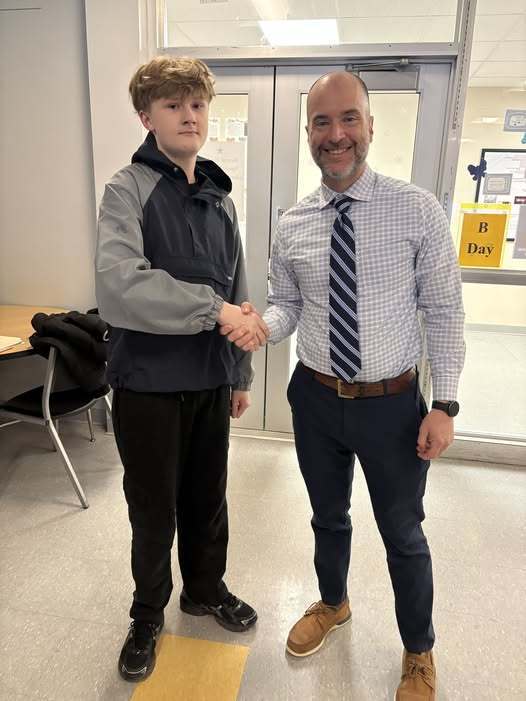 A boy in a gray and blue hoodie and man with a striped tie shaking hands.