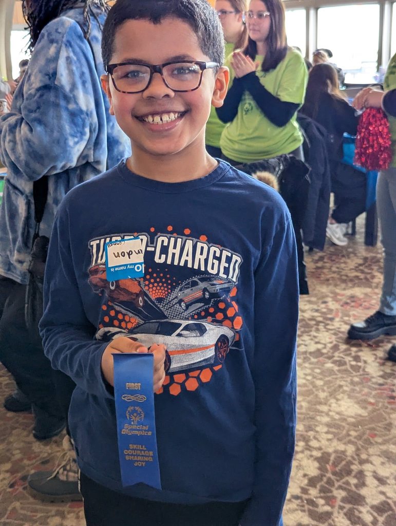 Young boy wearing a blue shirt with cars on it and holding his blue ribbon. 