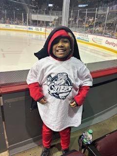 Young boy wearing a White Erie Otters shirt smiling in front of an ice rink.