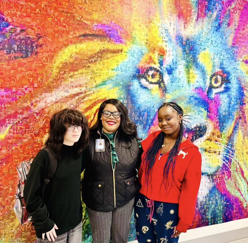 Students and women standing in front of a colorful piece of artwork on the wall.