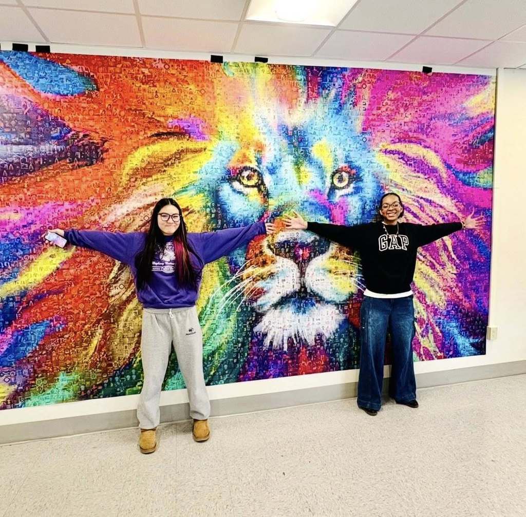 Students standing in front of a colorful piece of artwork on the wall and holding there hands open wide.