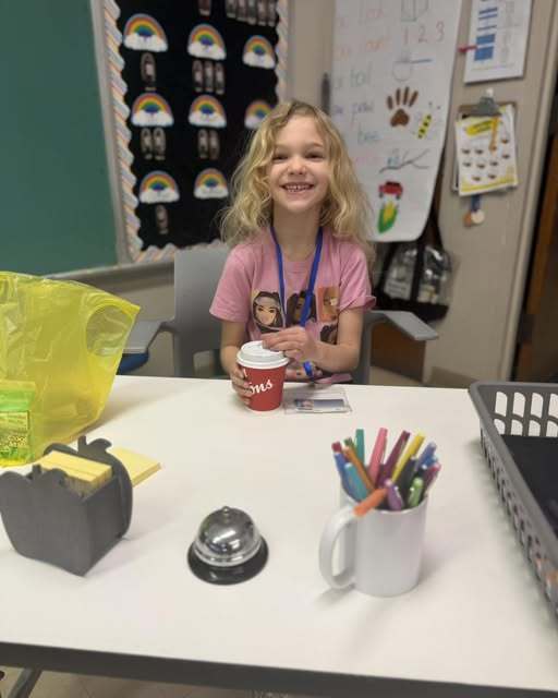 Young girl in a purple shirt smiling at a desk in school while she holds a cup of coffee from Tim Hortons. 