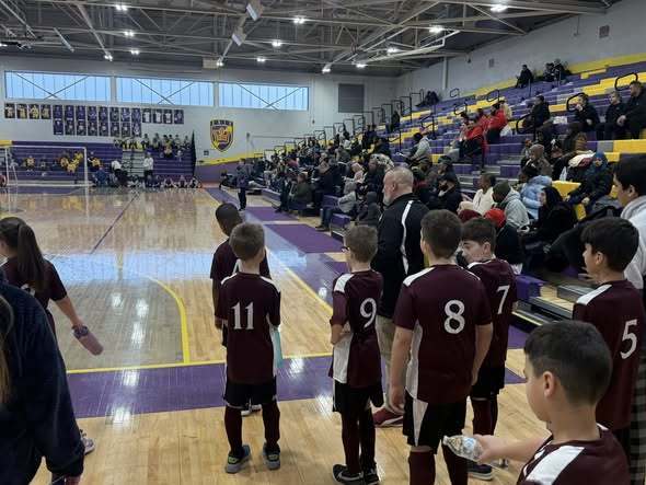 Students standing on a basketball court while audience sits in the bleachers and watches.