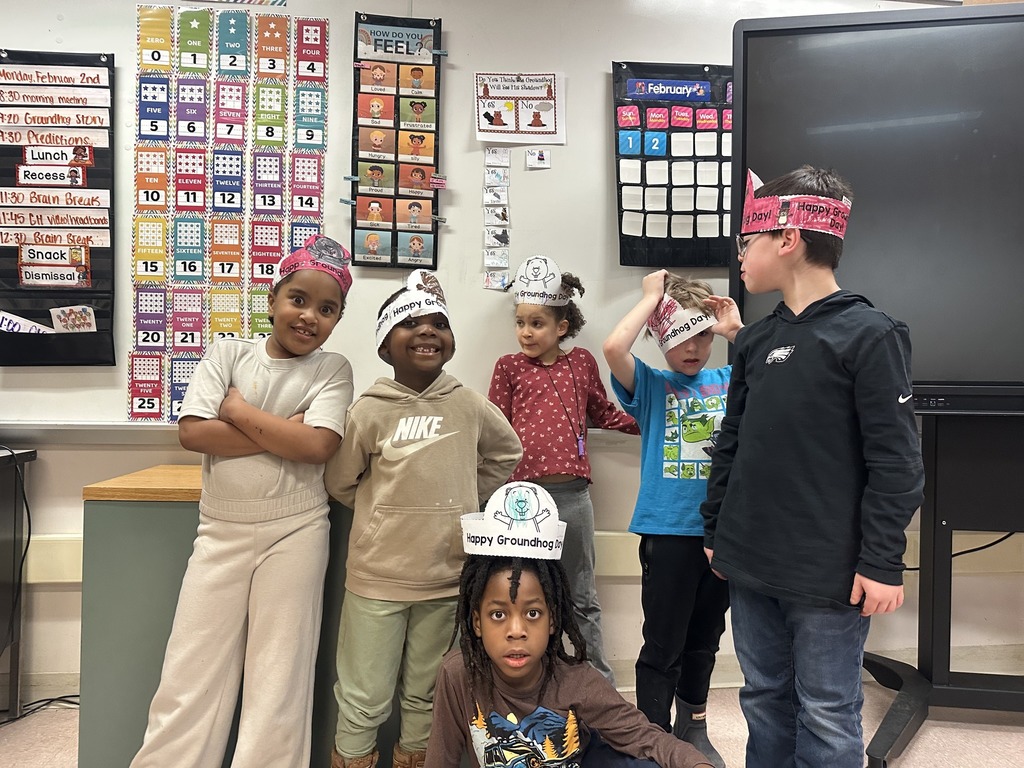 Kids in a classroom smiling and wearing paper headbands on their heads.