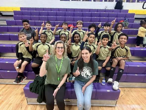 Group of kids and adults wearing green and  smiling while they point up and sit on the bleachers. 