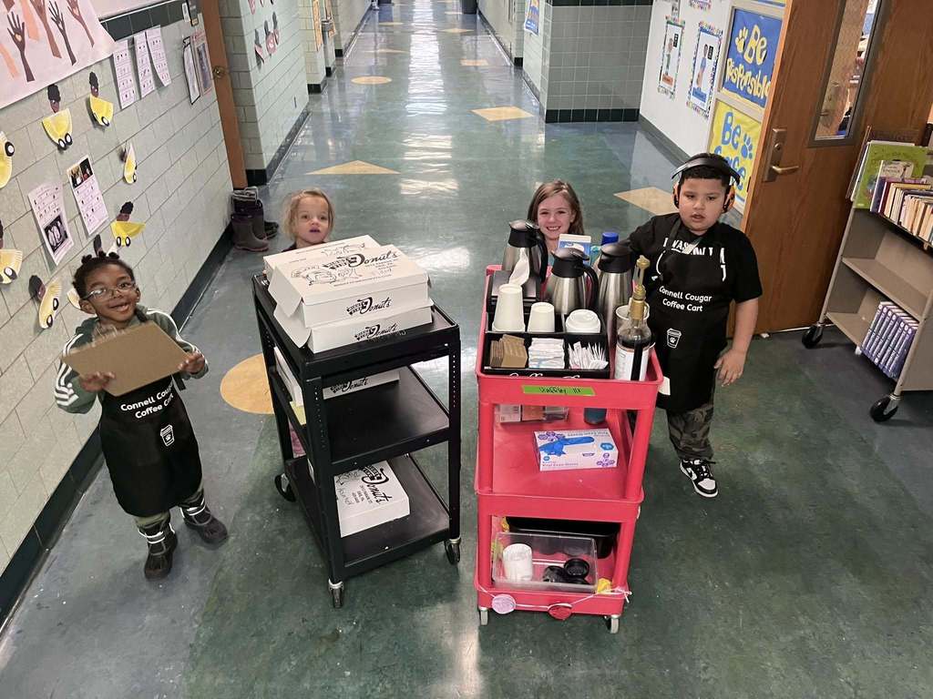 Kids smiling and they are standing behind a clipboard, a cart of donuts, and a cart for coffee. 