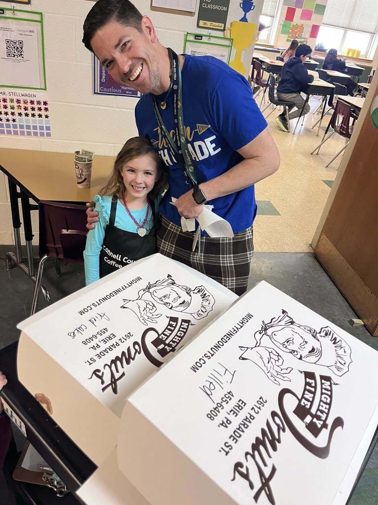 A young girl smiling next to a man in a blue shirt and there is boxes of donuts in front of them. 