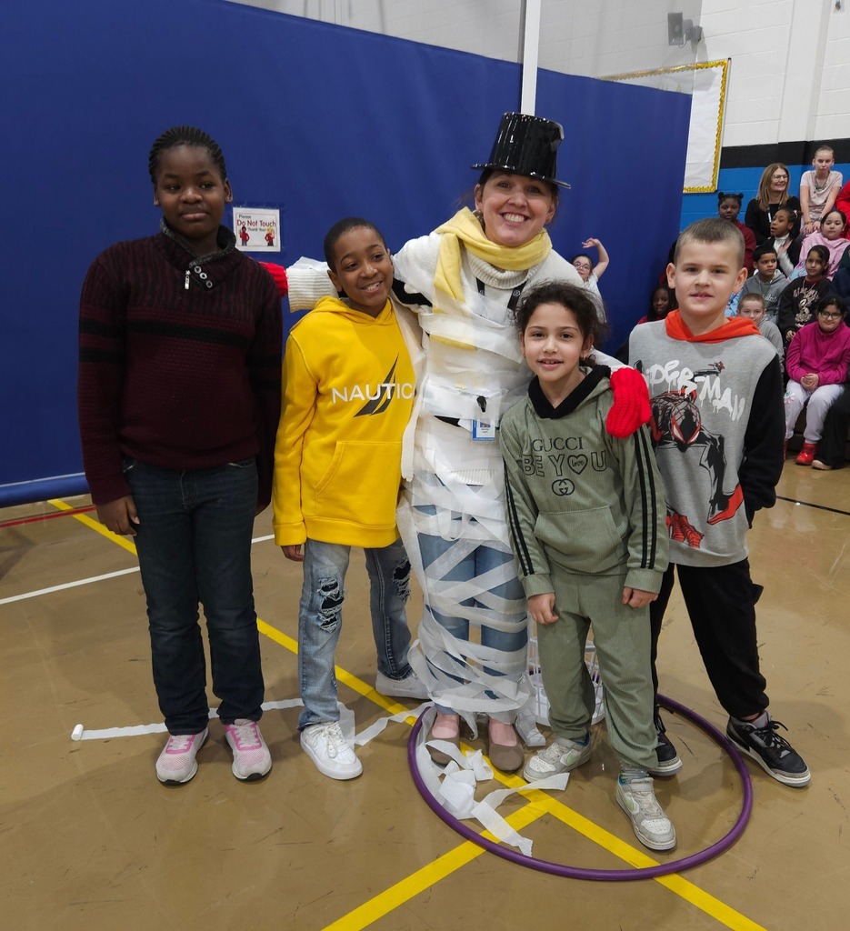 Students standing next to a women wearing a black top hat, a scarf , and is covered in toilet paper.