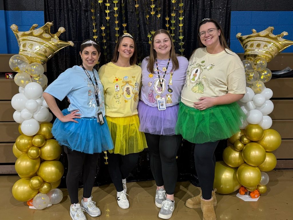 Four adult women dressed in tutus and shirts and they are all wearing crowns.