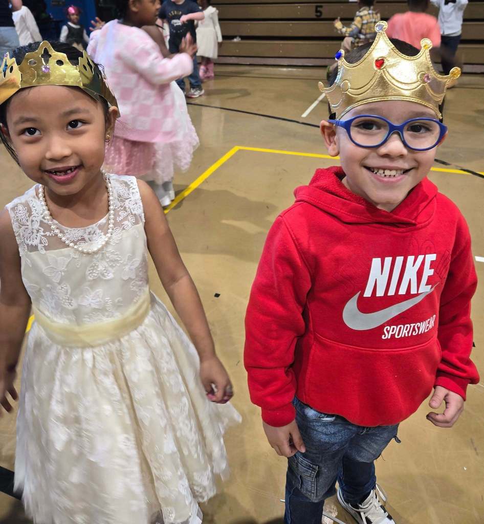 Young girl wearing a princess dress and crown is standing next to a boy wearing a red hoodie and a crown.