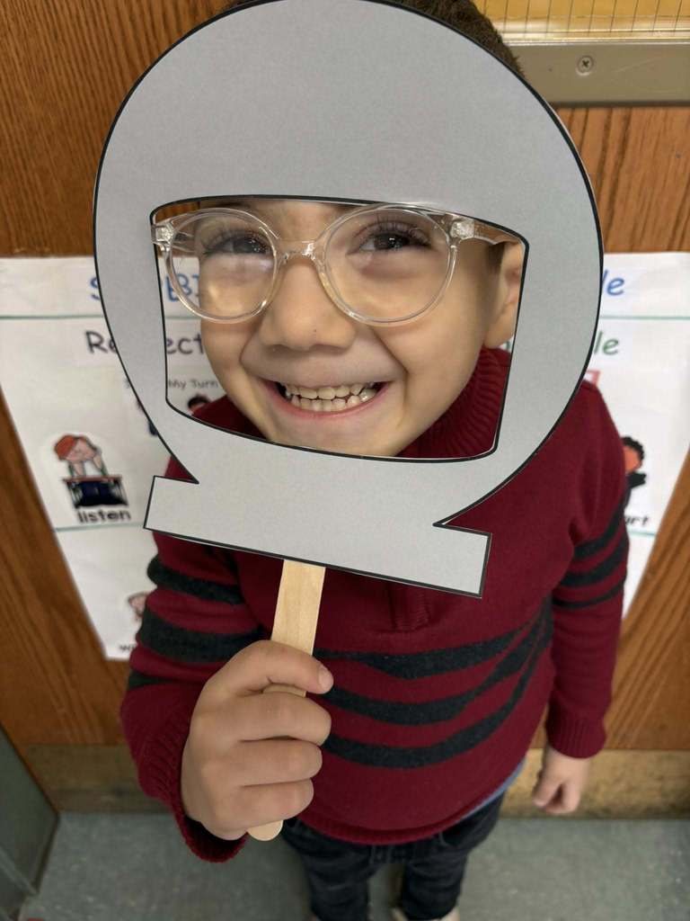Kid in a burgundy shirt with black stripes is holding up a cutout of a astronaut helmet on its face.