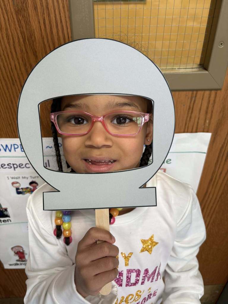 Kid in a white shirt is holding up a cutout of a astronaut helmet on its face.