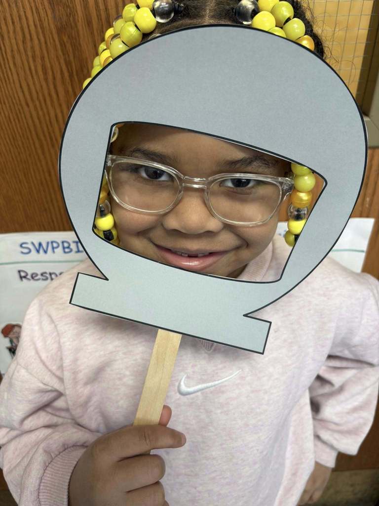 Kid in a pink shirt is holding up a cutout of a astronaut helmet on its face.