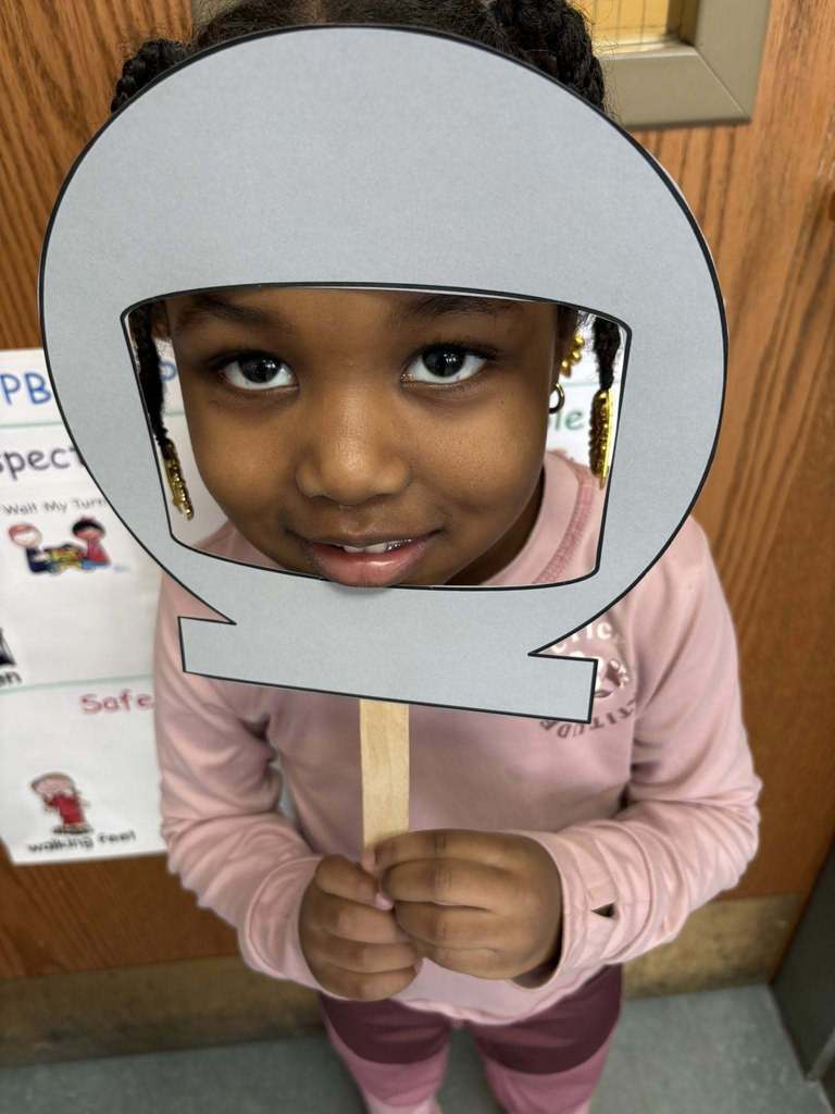 Kid in a pink shirt is holding up a cutout of a astronaut helmet on its face.