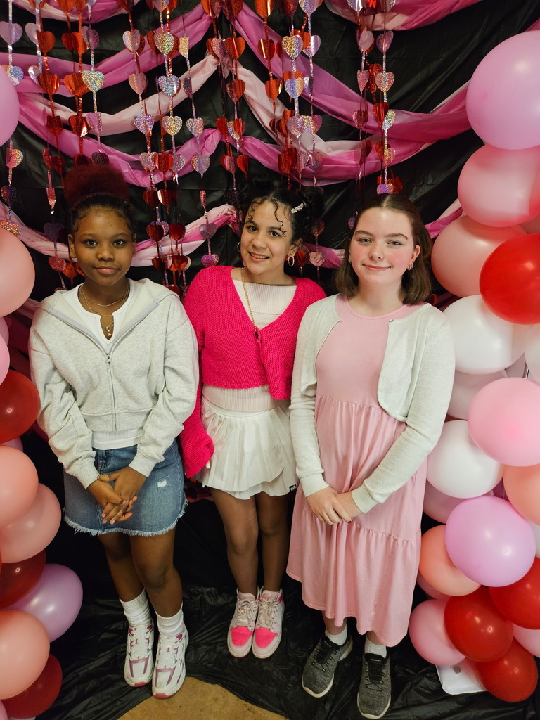 Three young girls standing in front of pink, red, and white decorations. 