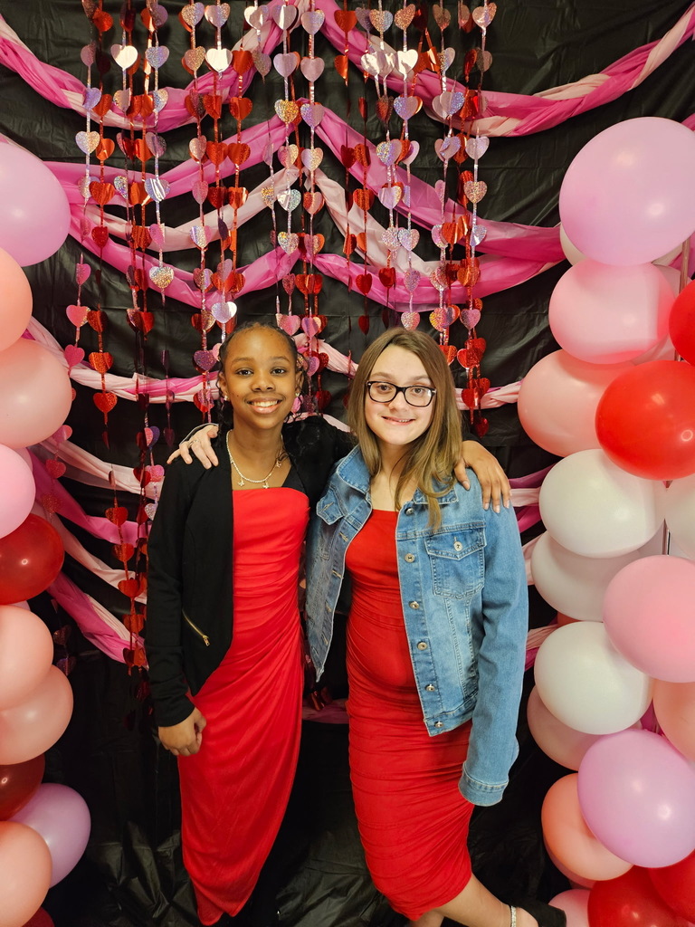 Two young girls hugging in front of pink, red, and white decorations. 