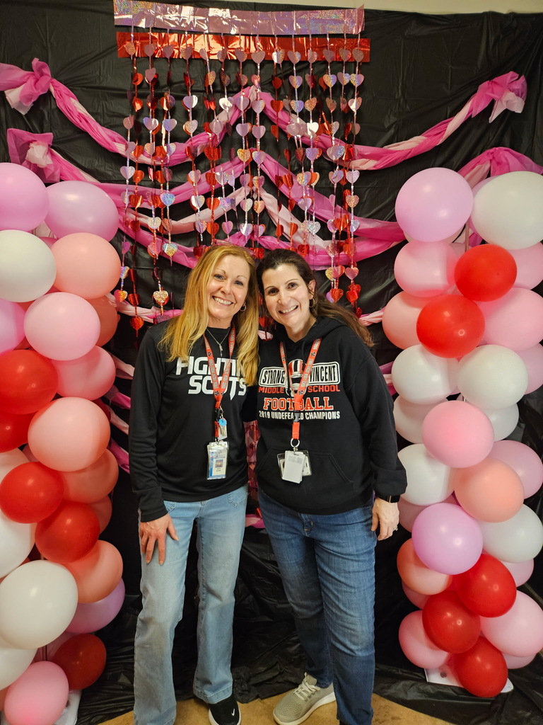 Two adult females in black tops standing in front of pink, red, and white decorations. 