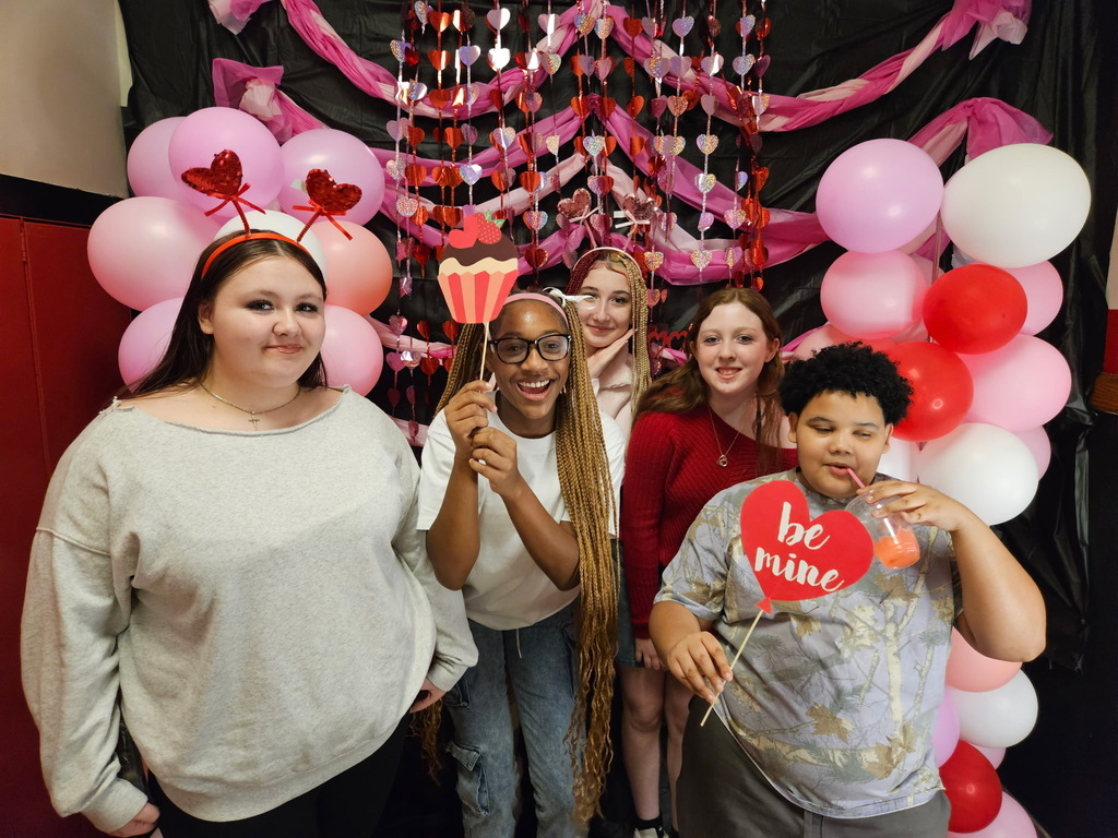 Group of kids holding props in front of pink, red, and white decorations. 