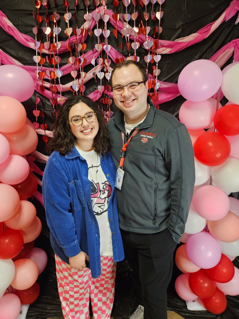 Two adults smiling in front of pink, red, and white decorations. 