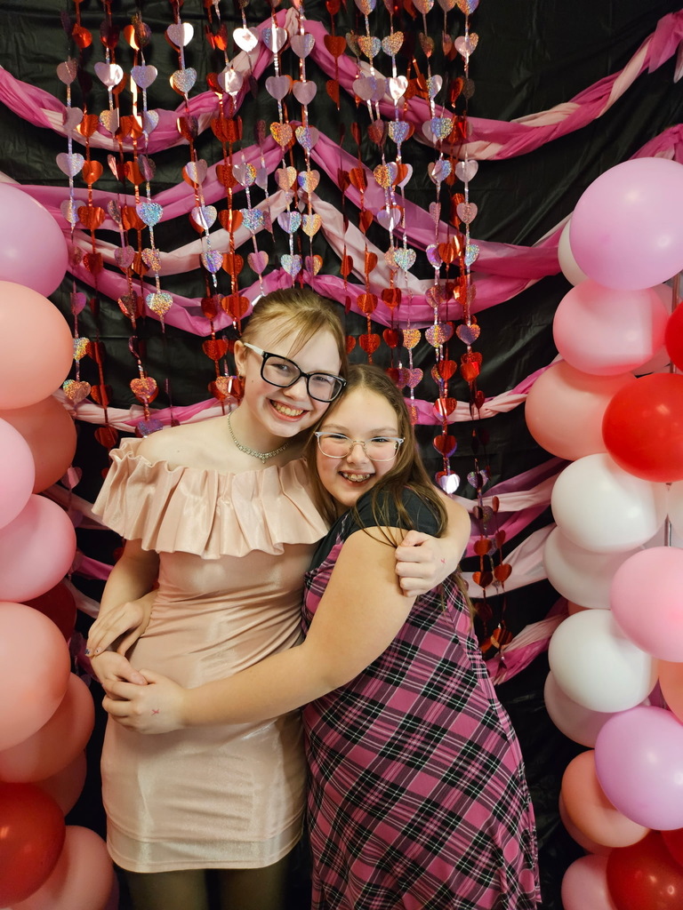 Two young girls hugging in front of pink, red, and white decorations. 