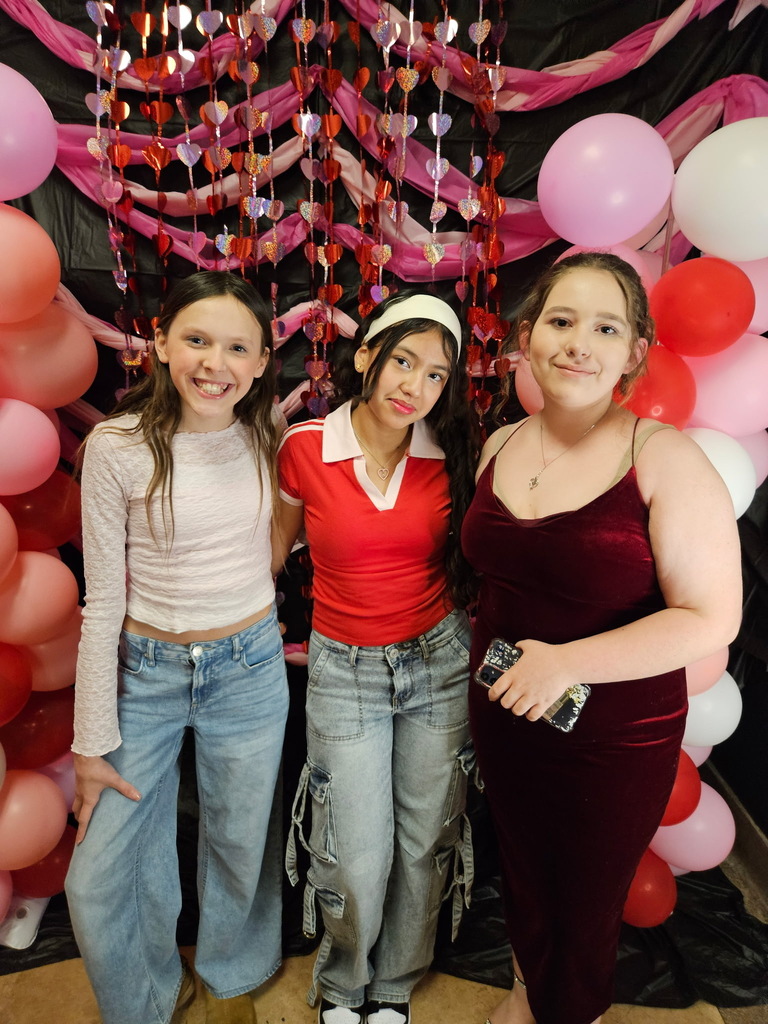 Three young girls standing in front of pink, red, and white decorations. 