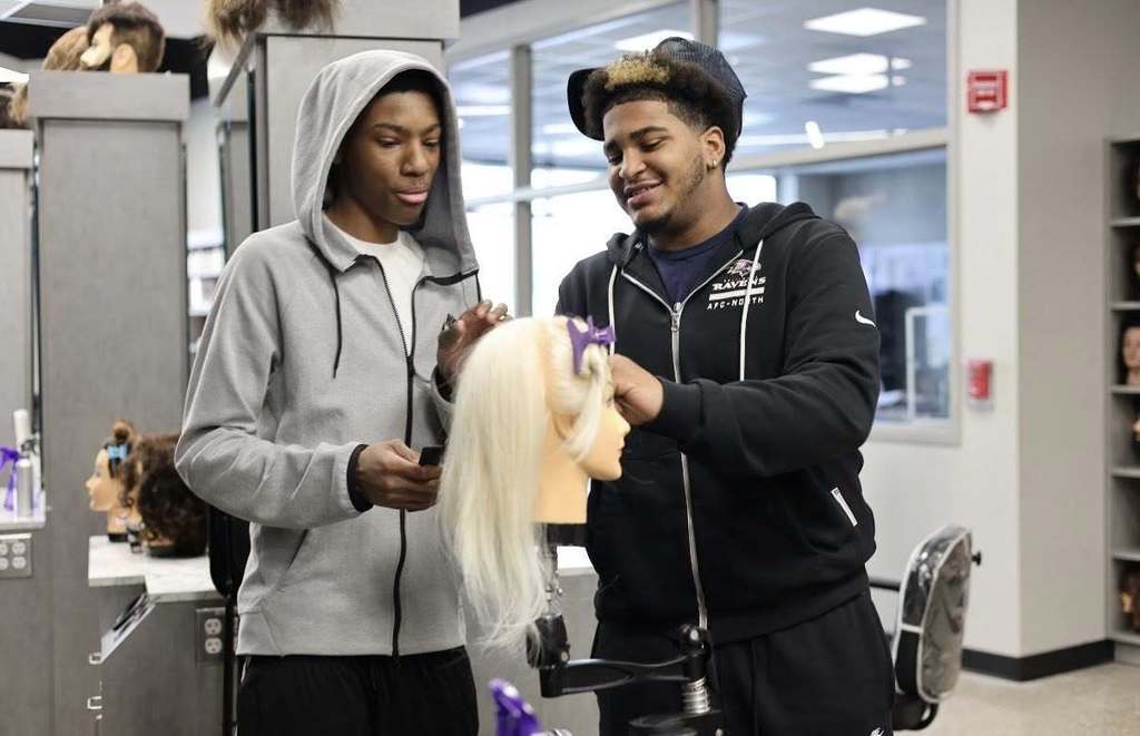 Two people stand around a mannequin head, possibly in a beauty school, with one holding the head and the other looking at it.