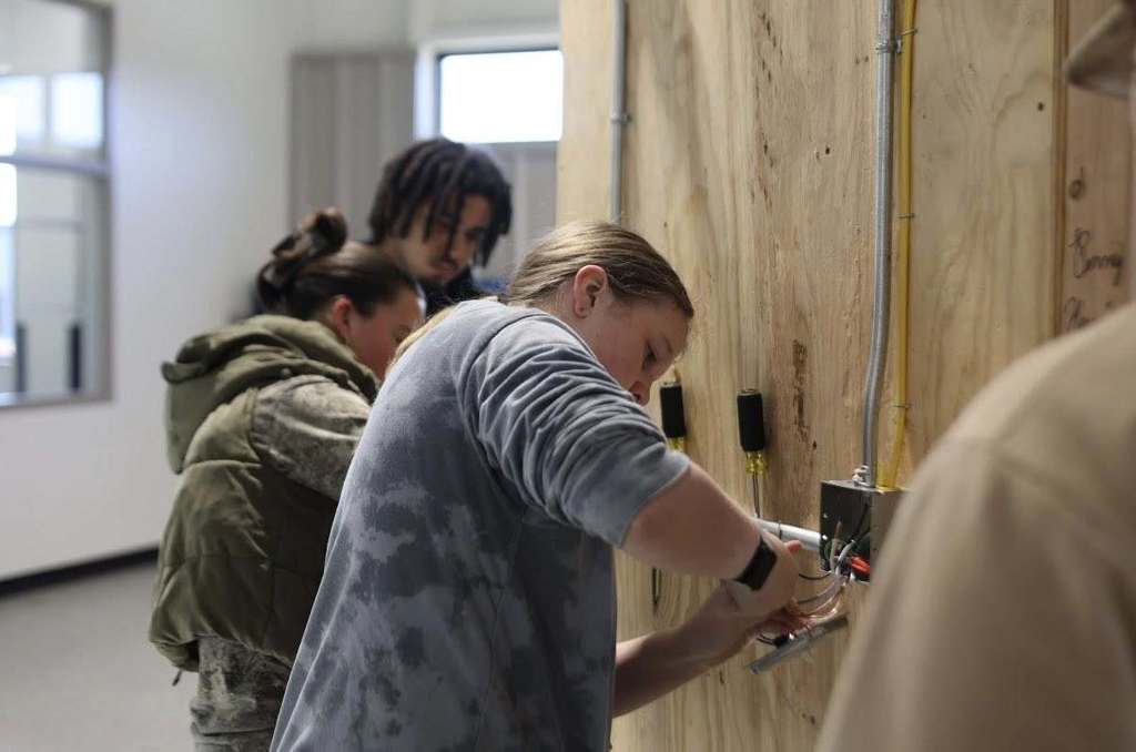 Four people work on an electrical box on a wooden wall, with a window and door in the background.