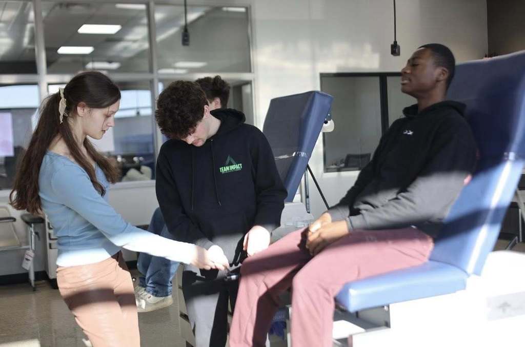 Three individuals in a clinic; one sits in a blue chair while another adjusts their leg. Background: a hospital room.