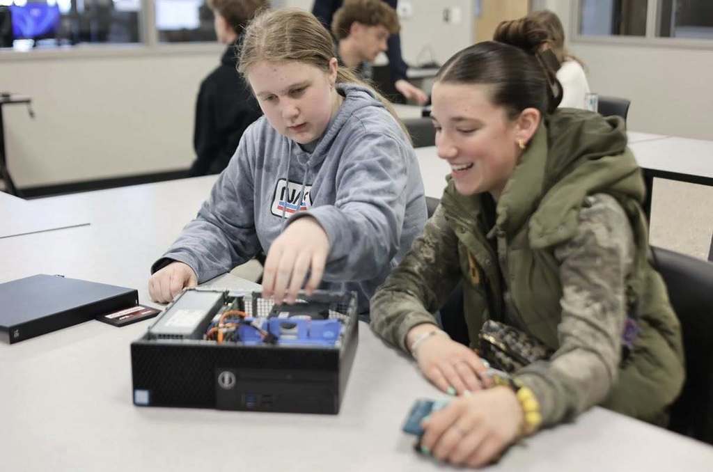 Two people sit at a desk, one wearing a blue hoodie, the other in a camouflage jacket. They focus on a computer box.