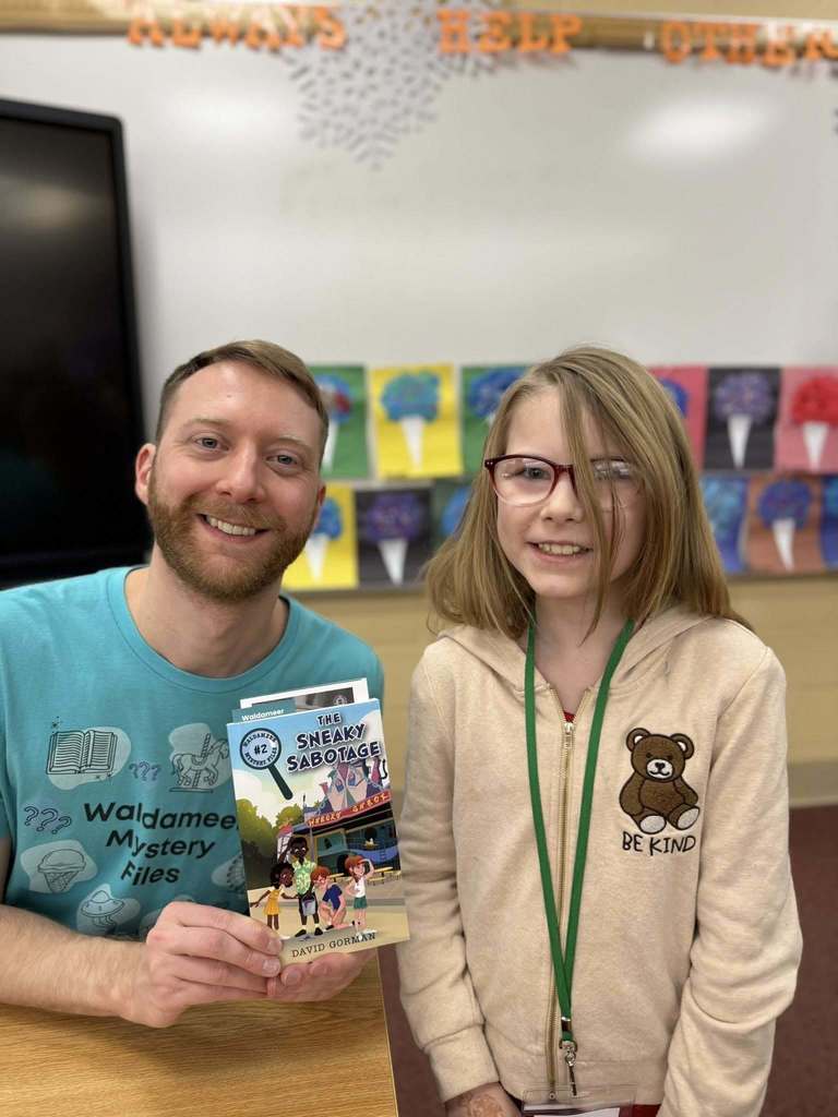 A man in a blue shirt is holding a book and smiling next to a kid in a tan hoodie.