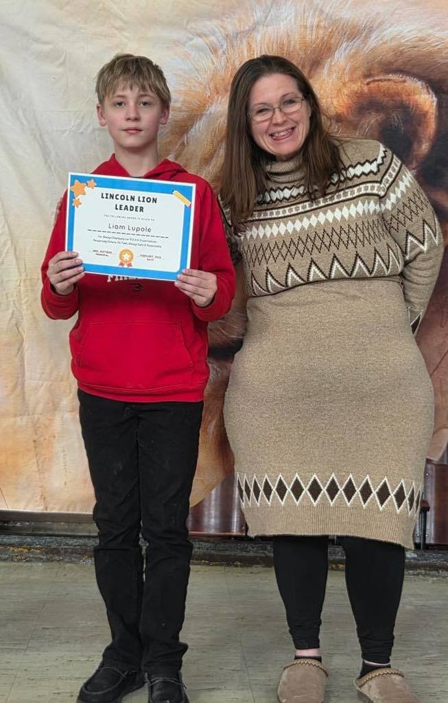 A women in a tan sweater dress smiling next to a kid  that is holding a certificate. 