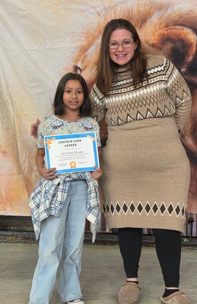 A women in a tan sweater dress smiling next to a kid  that is holding a certificate. 