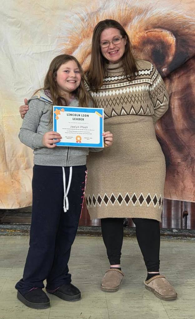A women in a tan sweater dress smiling next to a kid  that is holding a certificate. 
