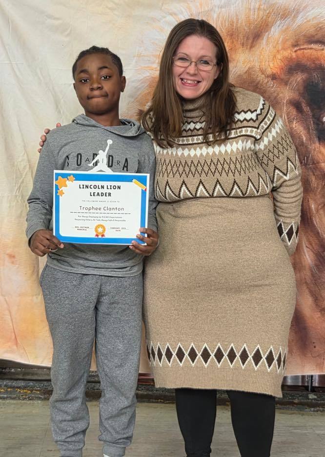A women in a tan sweater dress smiling next to a kid  that is holding a certificate. 