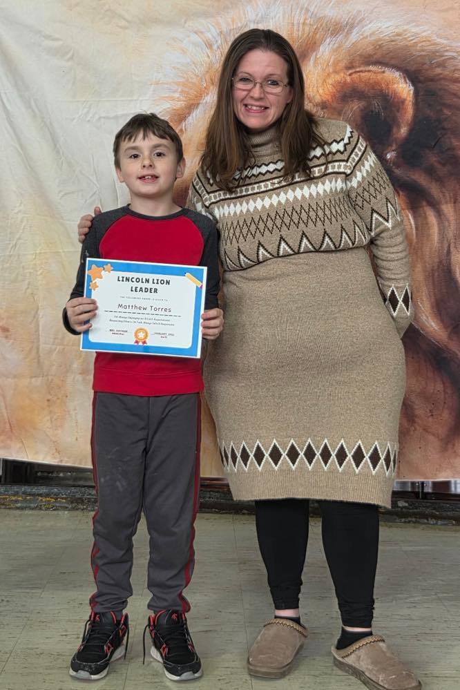 A women in a tan sweater dress smiling next to a kid  that is holding a certificate. 