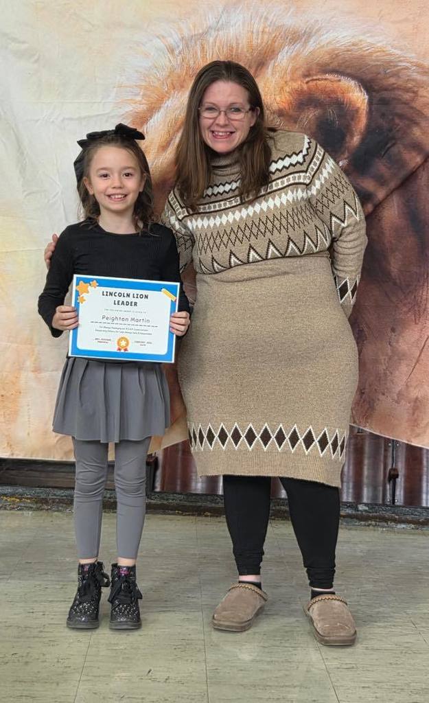 A women in a tan sweater dress smiling next to a kid  that is holding a certificate. 